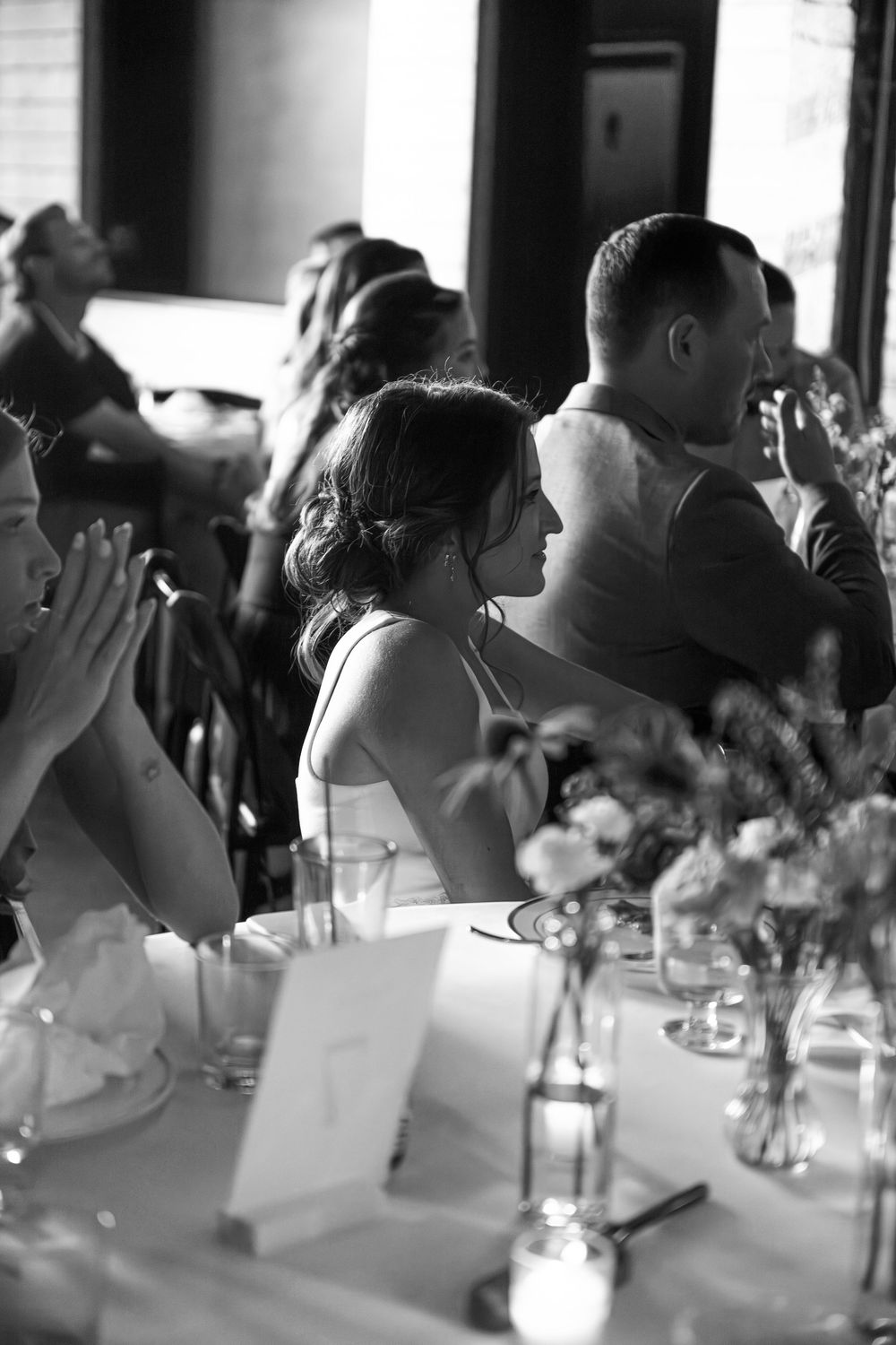 Black and white photo of wedding guests seated at reception tables with floral centerpieces and glassware.