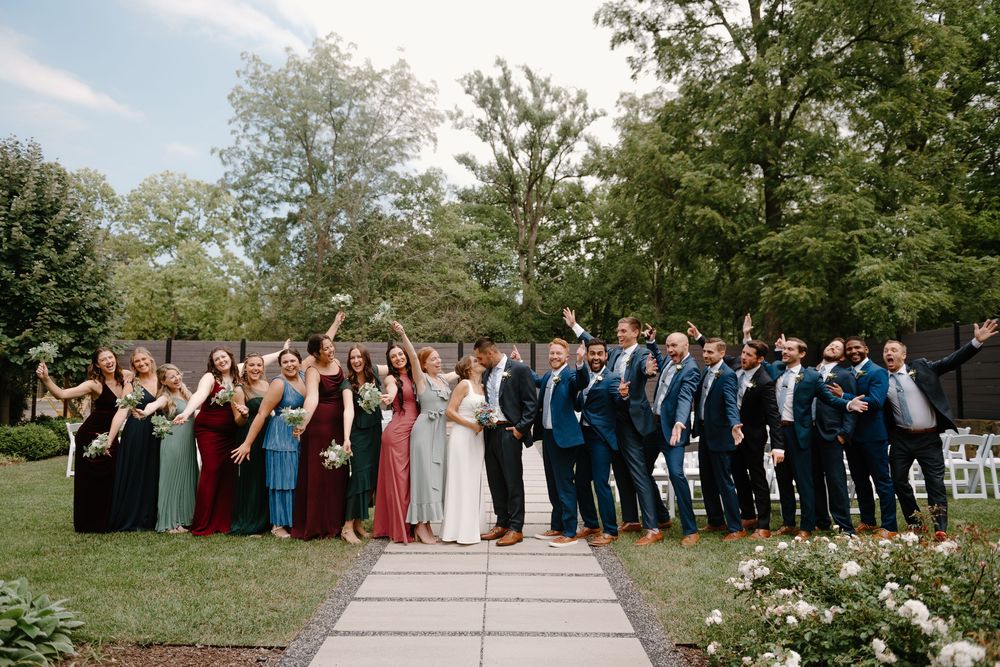 A wedding party in formal attire poses together on a garden path surrounded by greenery on a sunny day.