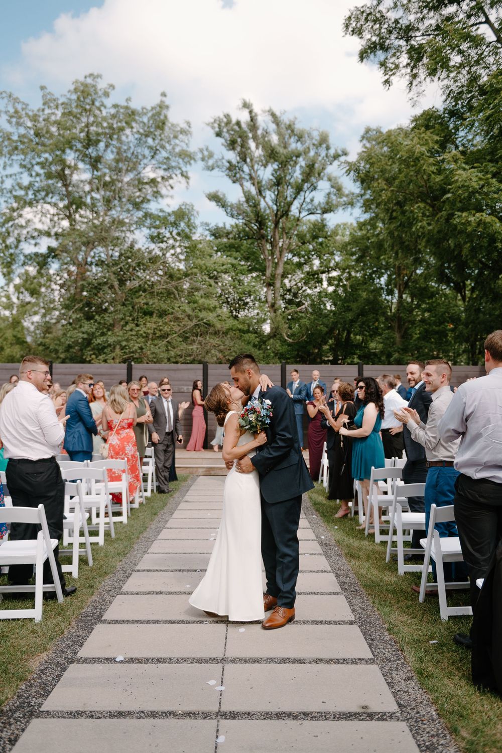Wedding ceremony guests stand and celebrate as newlyweds walk down outdoor aisle lined with white chairs and trees.
