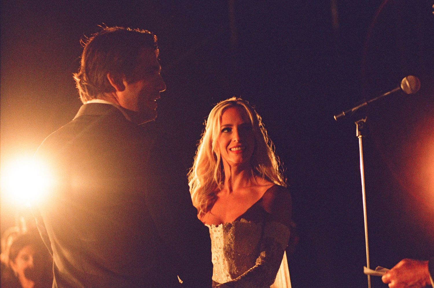 Performers on stage during a concert with dramatic orange lighting and a microphone stand visible.