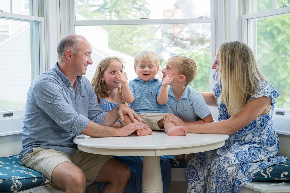 A family shares an intimate moment of laughter and conversation while gathered around a white table near bright windows.