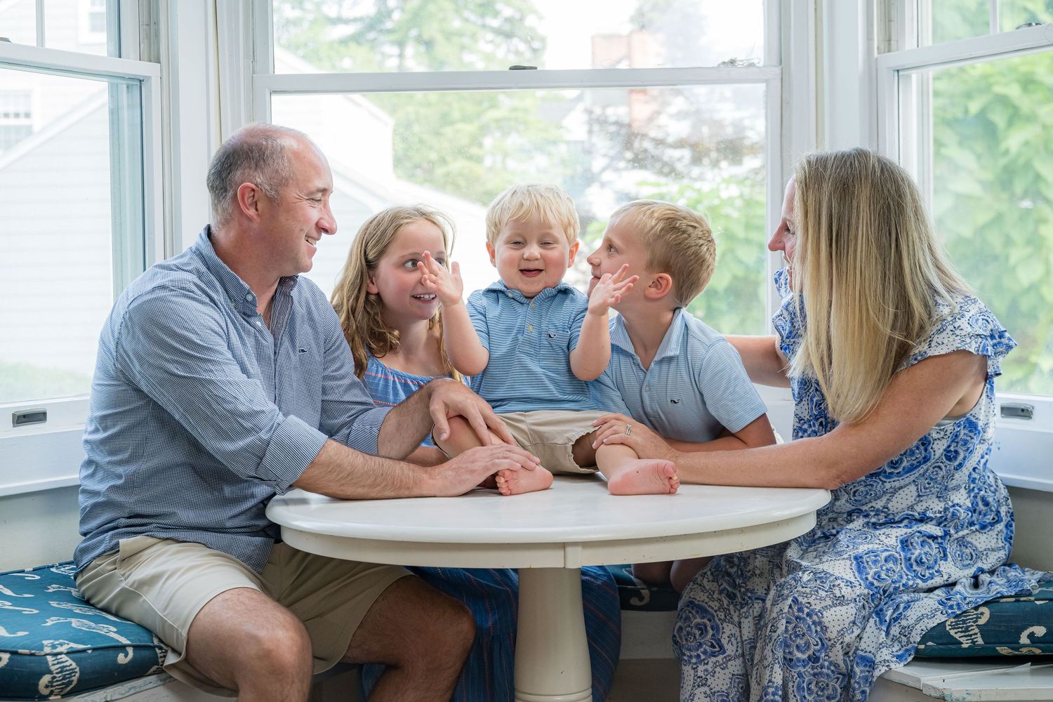 A family shares an intimate moment of laughter and conversation while gathered around a white table near bright windows.