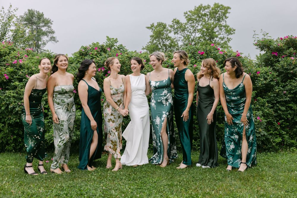 A group of women in emerald green floral and solid dresses stand together on grass surrounded by greenery.