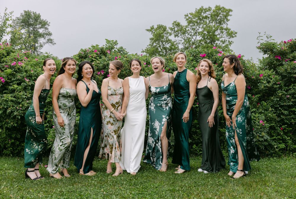 Bridesmaids pose together on grass wearing elegant emerald green and floral sequined gowns at outdoor wedding.