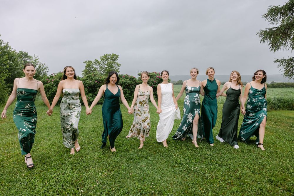 Wedding party in green and white dresses jump together in a playful sequence on a grassy field.