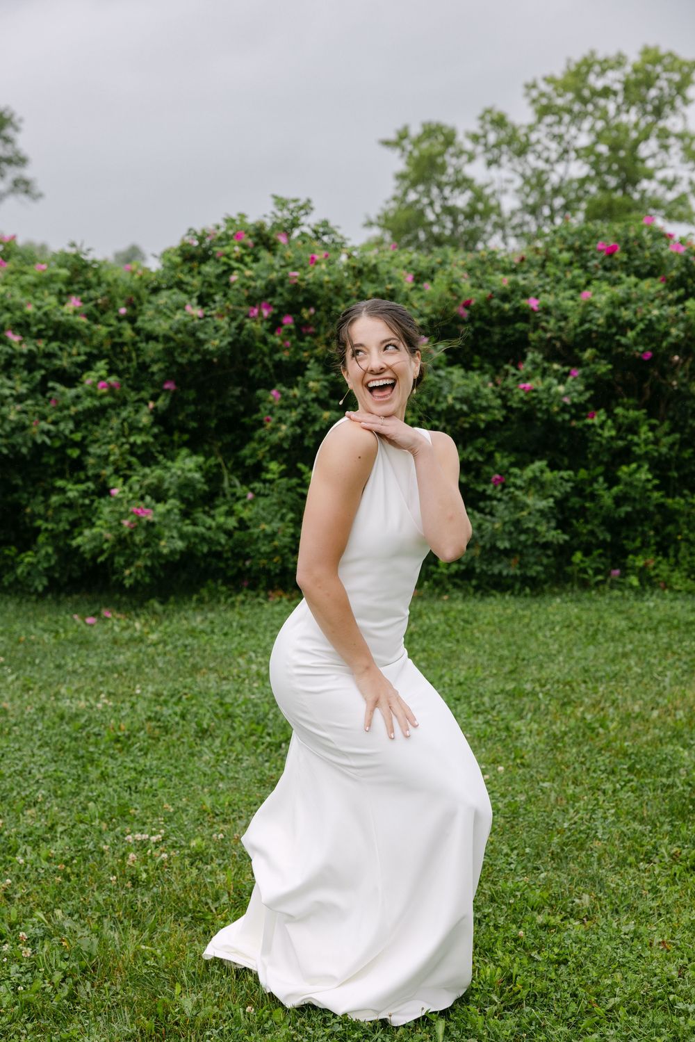 A joyful bride in a sleek white wedding dress poses playfully on grass with rose bushes in the background.