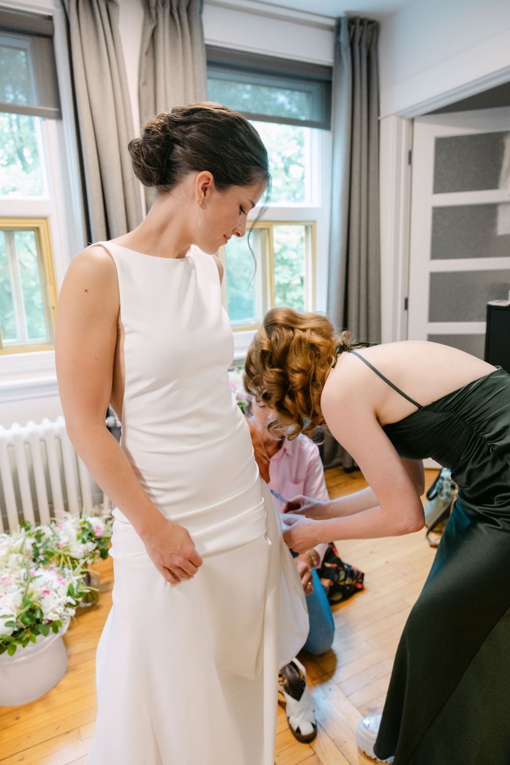 Helper adjusts a long white wedding dress during preparations in a sunlit room with hardwood floors and cream curtains.