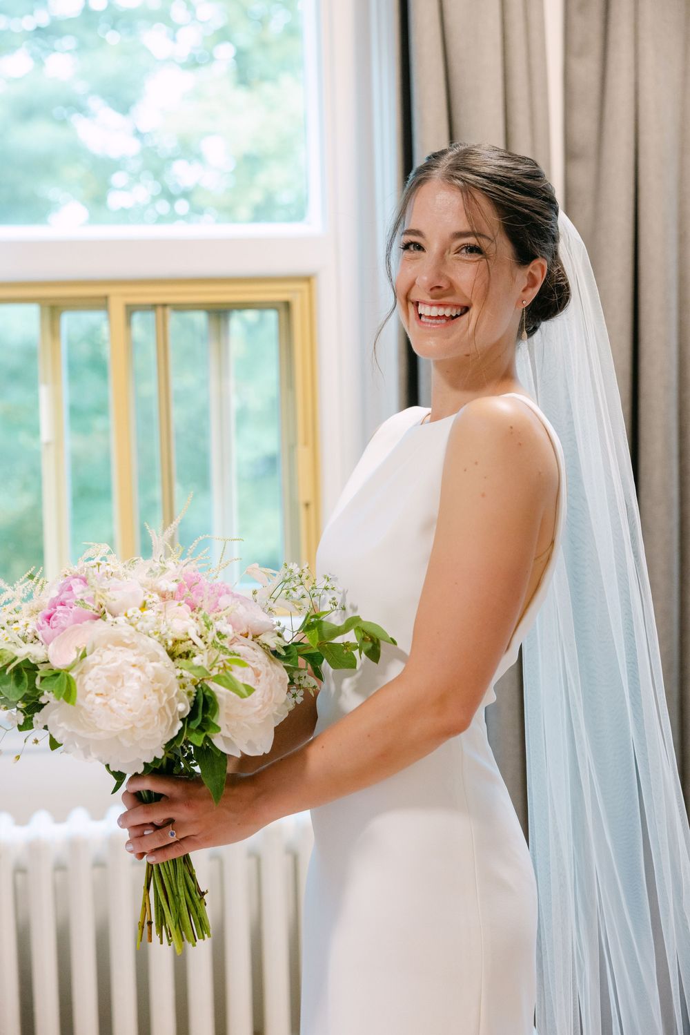 A bride in a white wedding dress and veil holds a delicate bouquet of white and pink flowers while posing in a bright room.