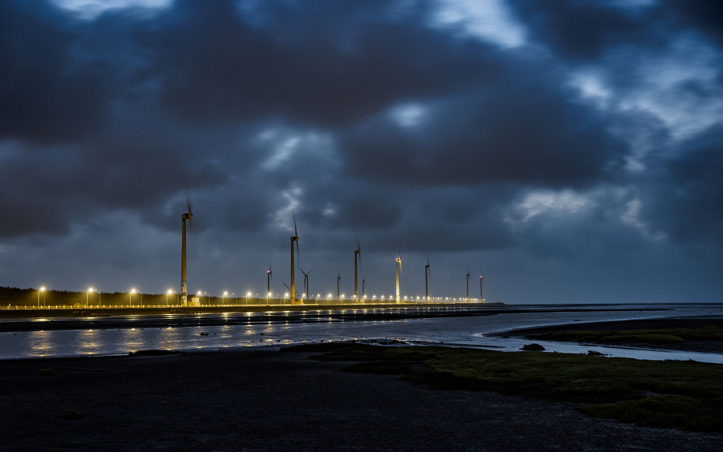Illuminated pier with wind turbines extends into dark waters under stormy skies.