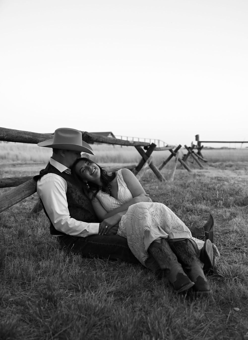 A romantic western-themed couple rests against a wooden fence in a rustic black and white photograph.