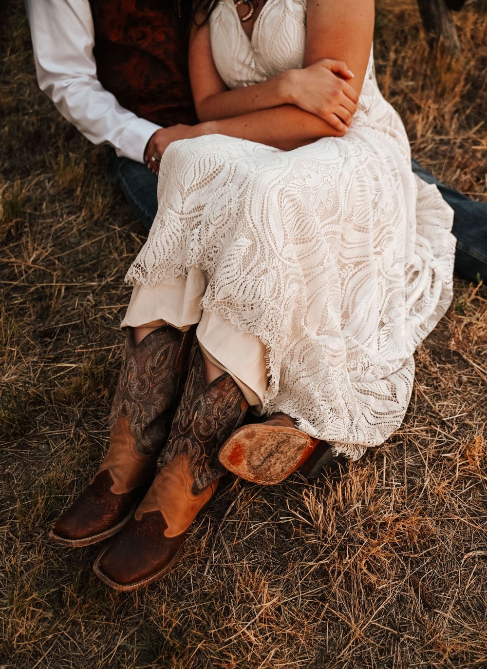 Close-up of lace wedding dress and brown leather boots in rustic outdoor setting.