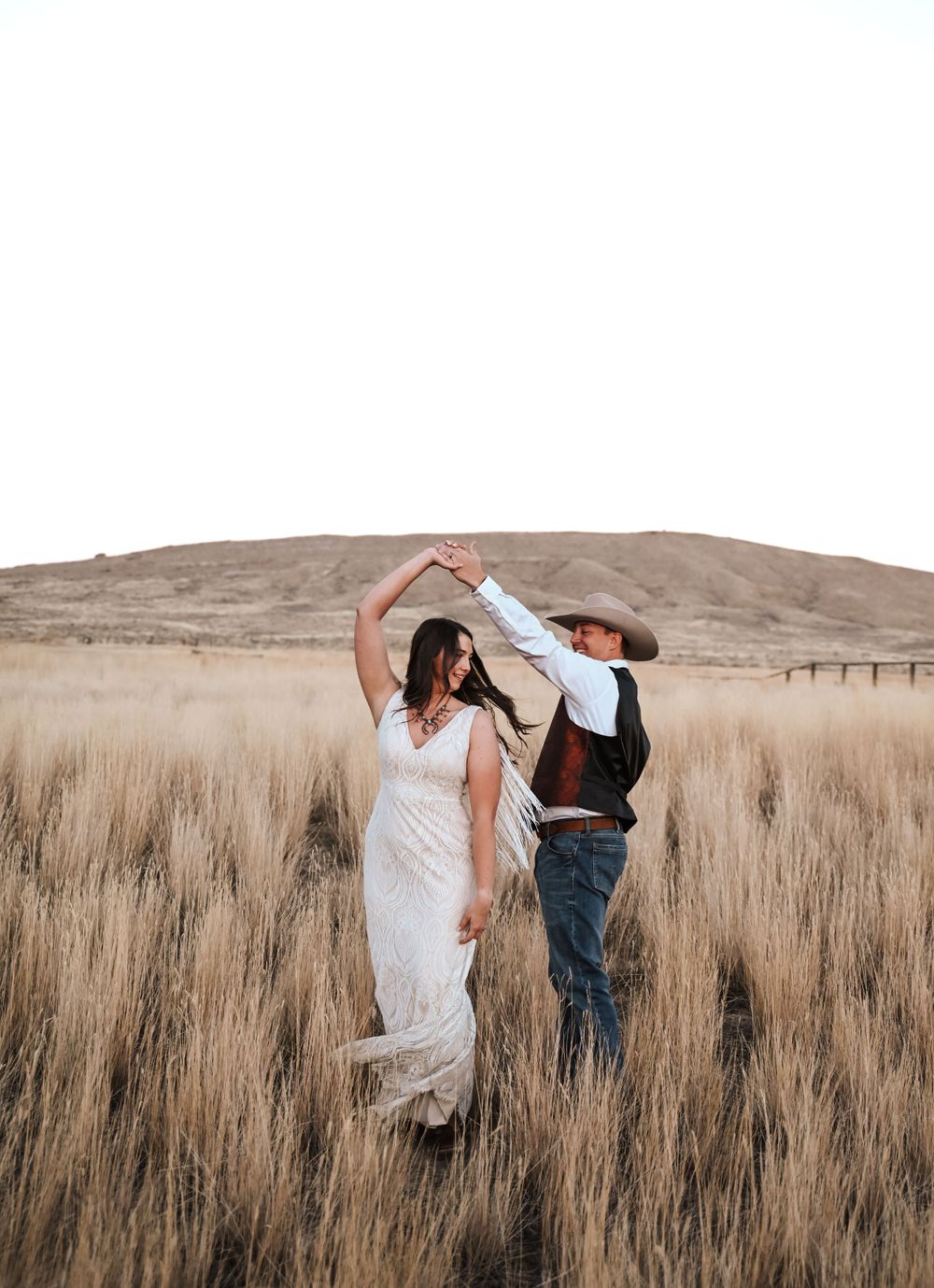 Couple dancing in tall grass field with mountains in background at sunset.