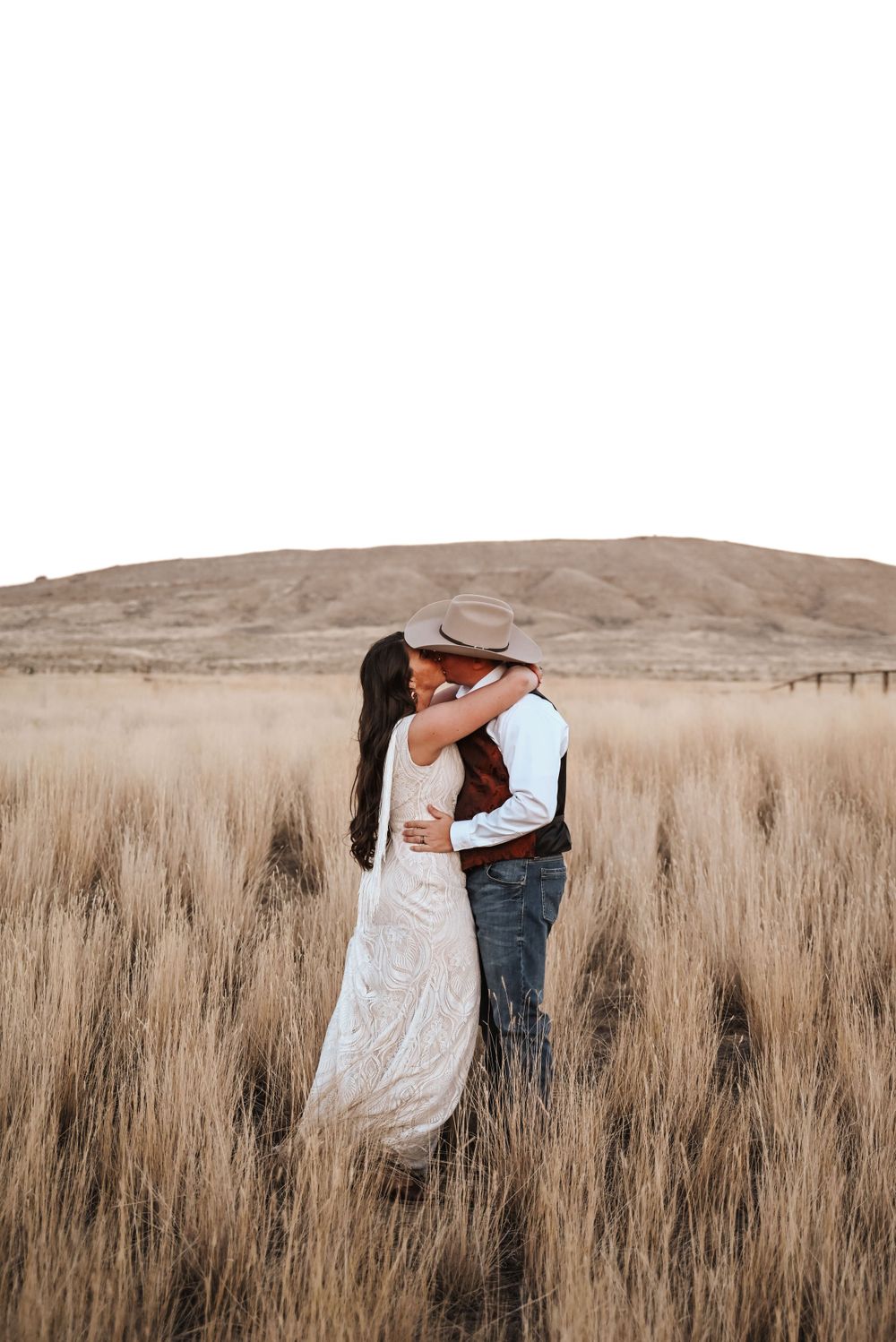 A romantic embrace between a couple in western attire standing in a golden wheat field at sunset.
