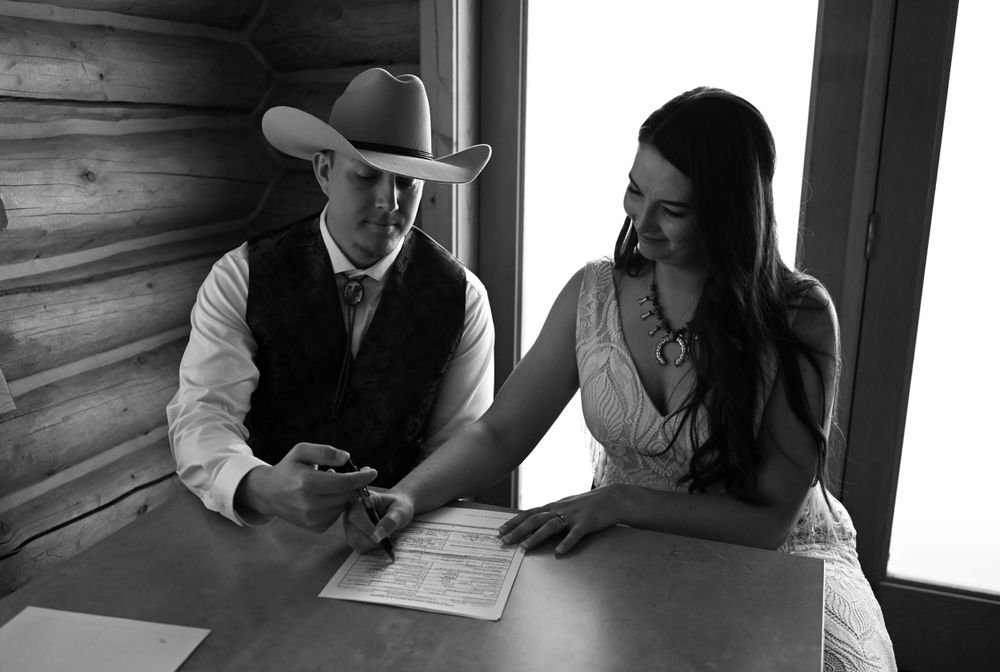 A couple in western attire signs papers at a wooden table in a rustic cabin.