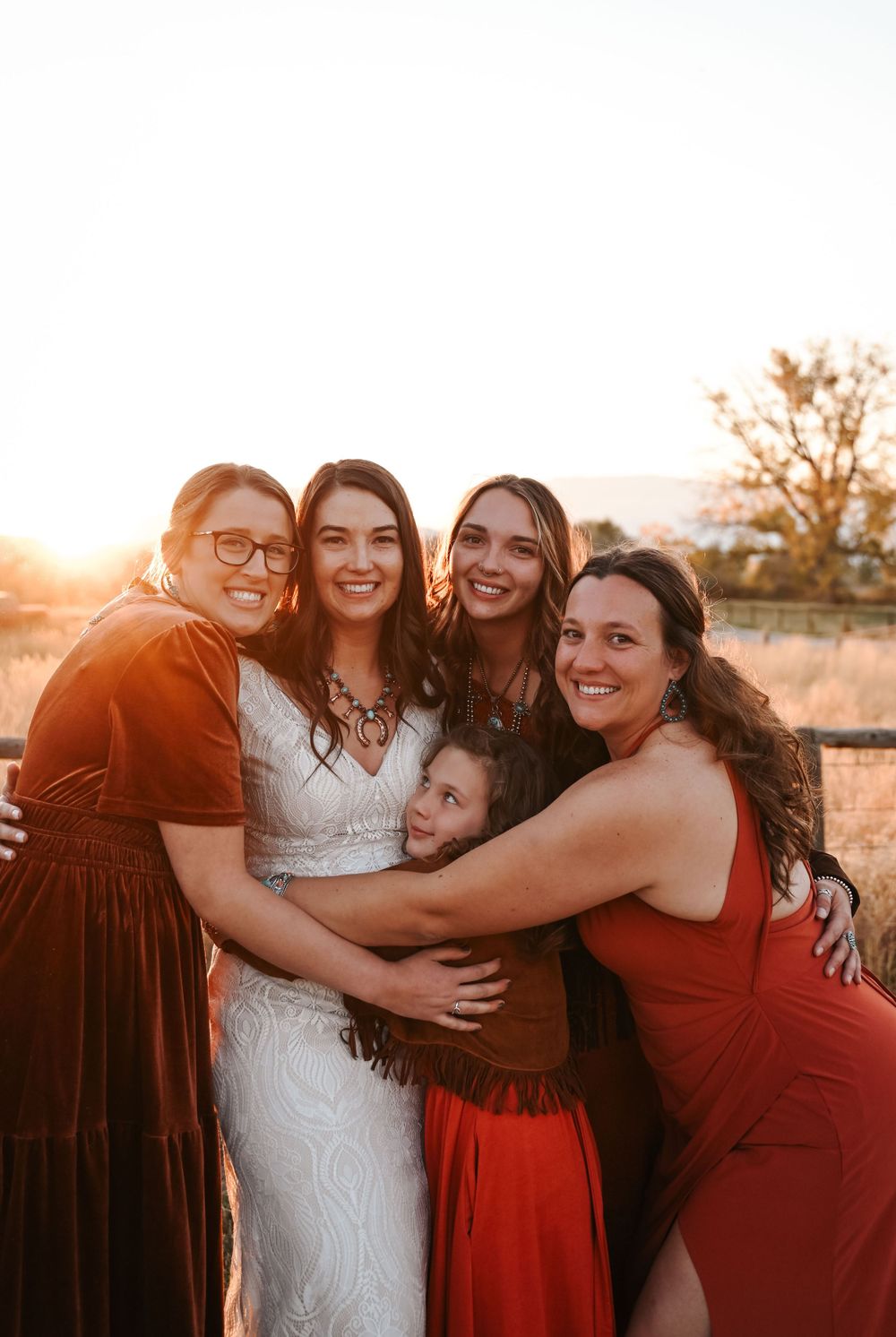 A joyful group hug between women in formal dresses at sunset.