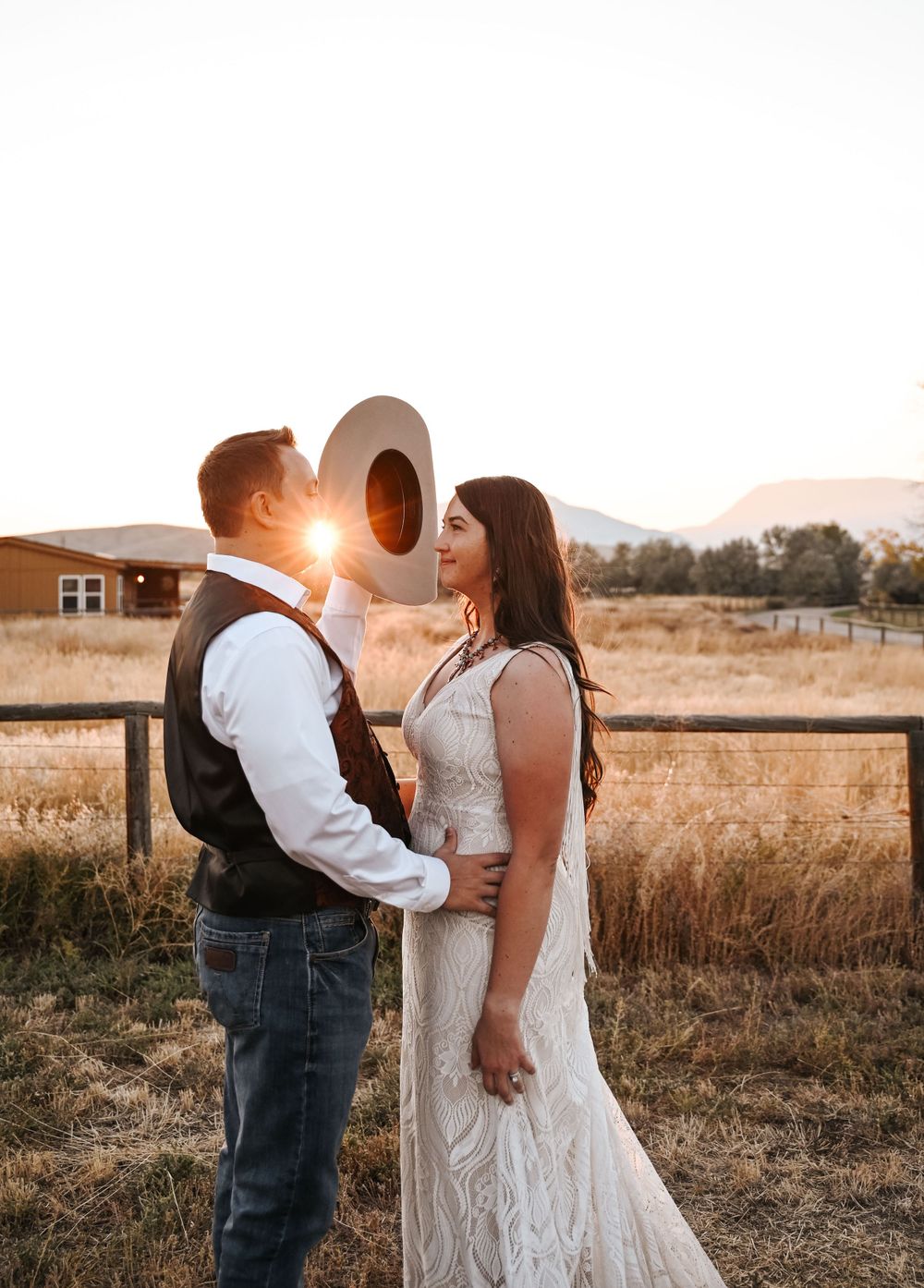 A romantic sunset wedding photo of a couple in western attire standing in a golden field.