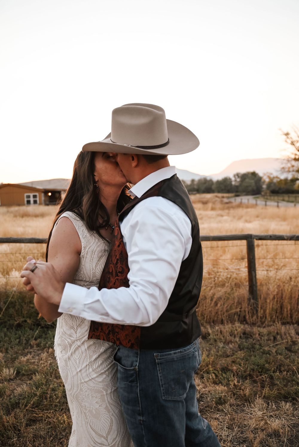 Intimate moment between newlyweds in country western attire by a rustic fence.