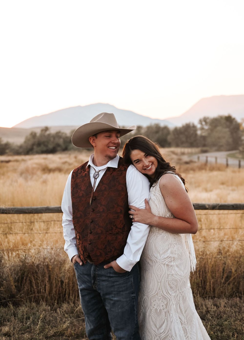 Newlyweds in rustic western style embrace against a mountain backdrop at sunset.