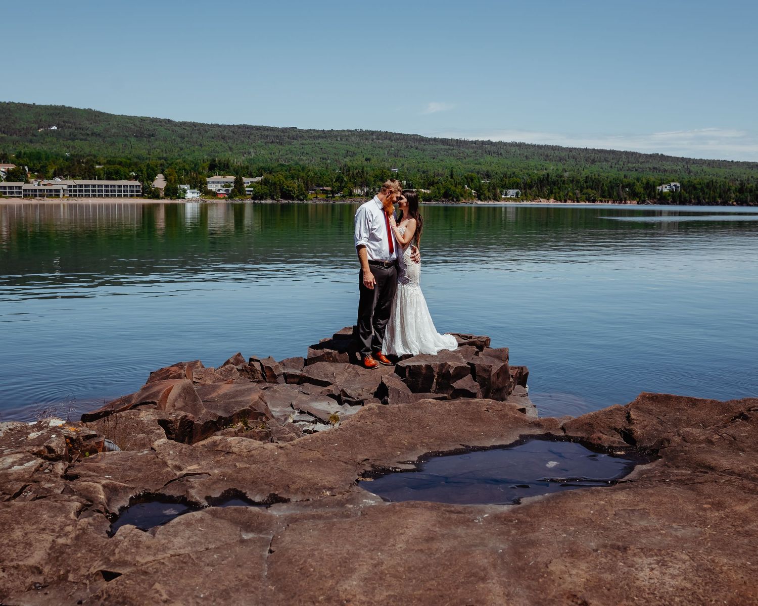 A couple poses on rocky outcrop by calm lake waters with trees in background.