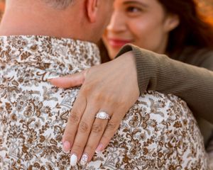 A hand displaying an engagement ring rests on a floral patterned shirt in warm lighting.