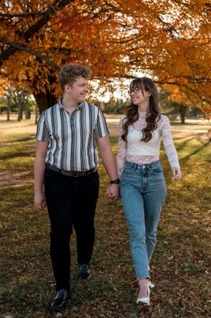 A couple strolls together on an autumn path beneath orange-colored trees and falling leaves engagement photoshoot.