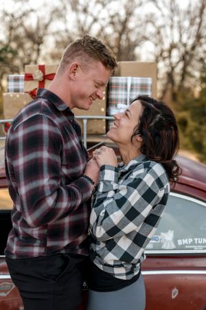 Couple laughing together in plaid shirts by vintage car with wrapped presents.
