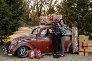 Couple poses with vintage red Volkswagen Beetle surrounded by wrapped Christmas presents.