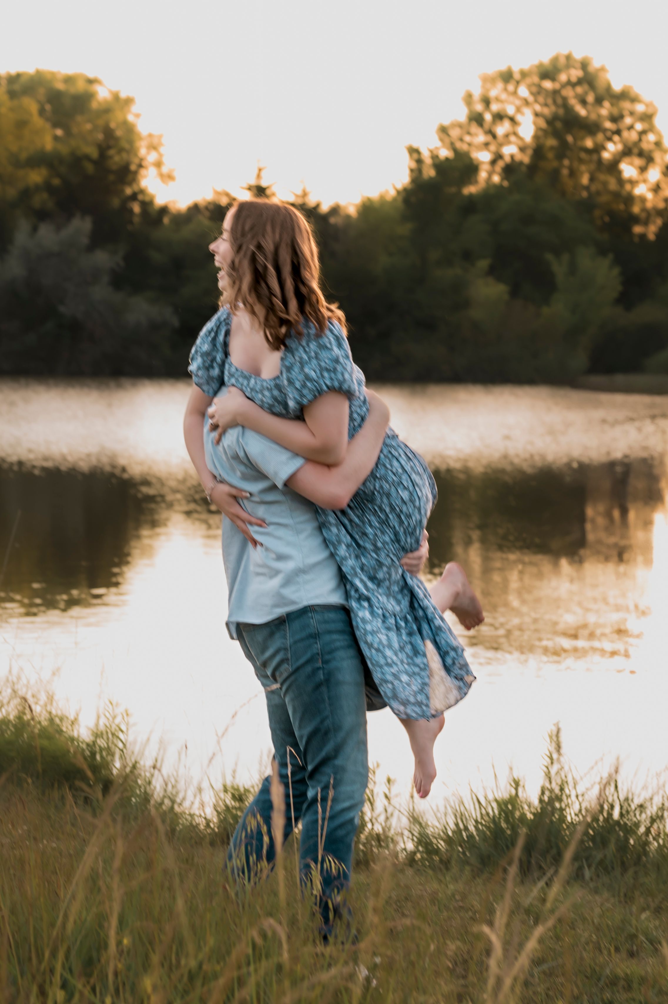 A romantic sunset photoshoot shows a couple embracing near a tranquil lake with tall grasses in the foreground.