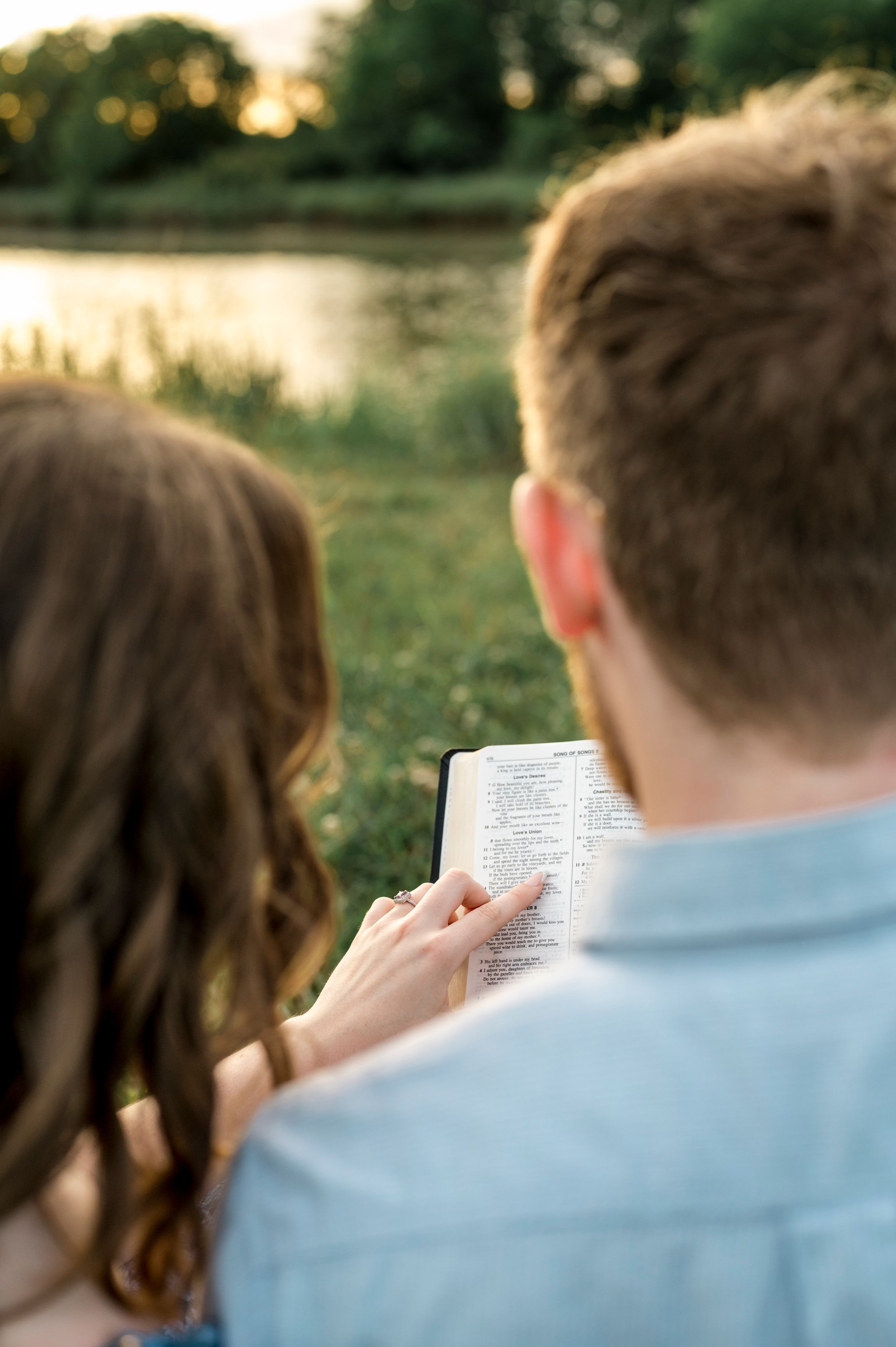 A couple reading from a book together by a tranquil lake during sunset.