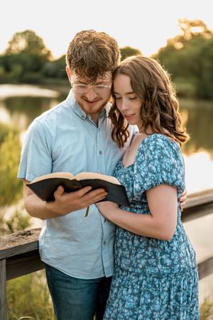 A couple sits close together reading a book on a wooden dock by a serene lake at sunset.