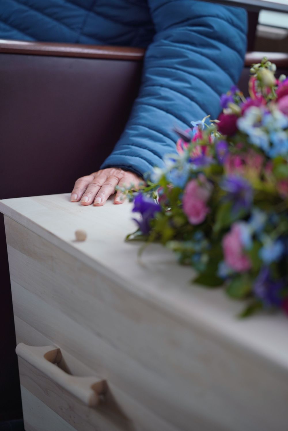 A hand rests on a white coffin adorned with colorful pink and purple funeral flowers.