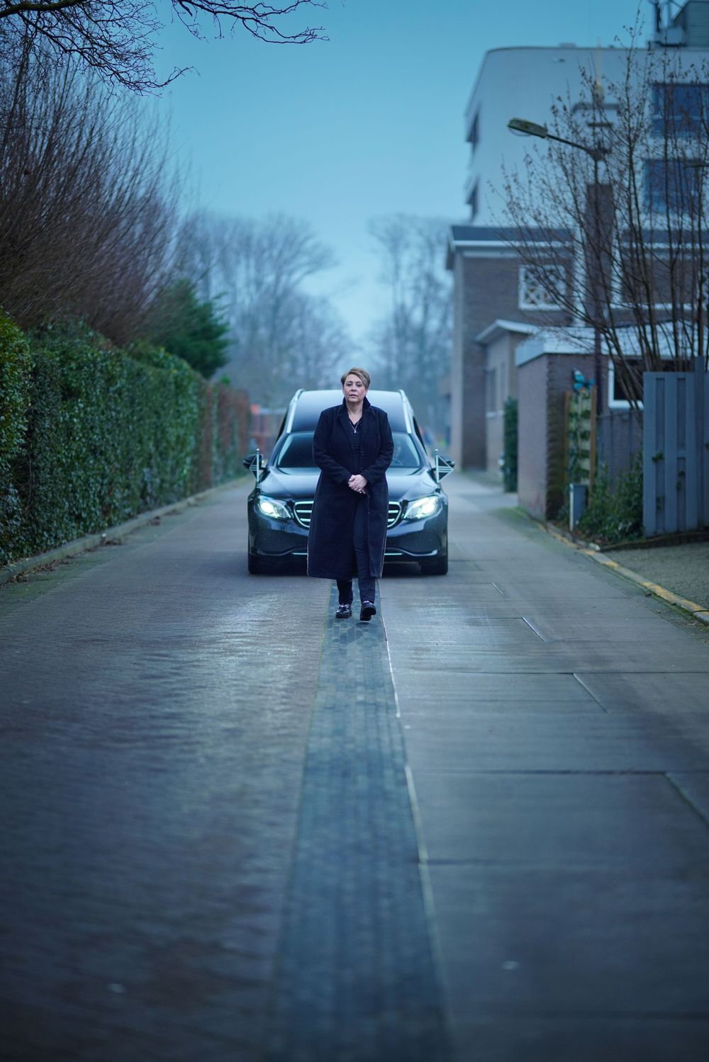 A person in dark clothing stands on a wet street in front of a parked BMW on a moody winter day.