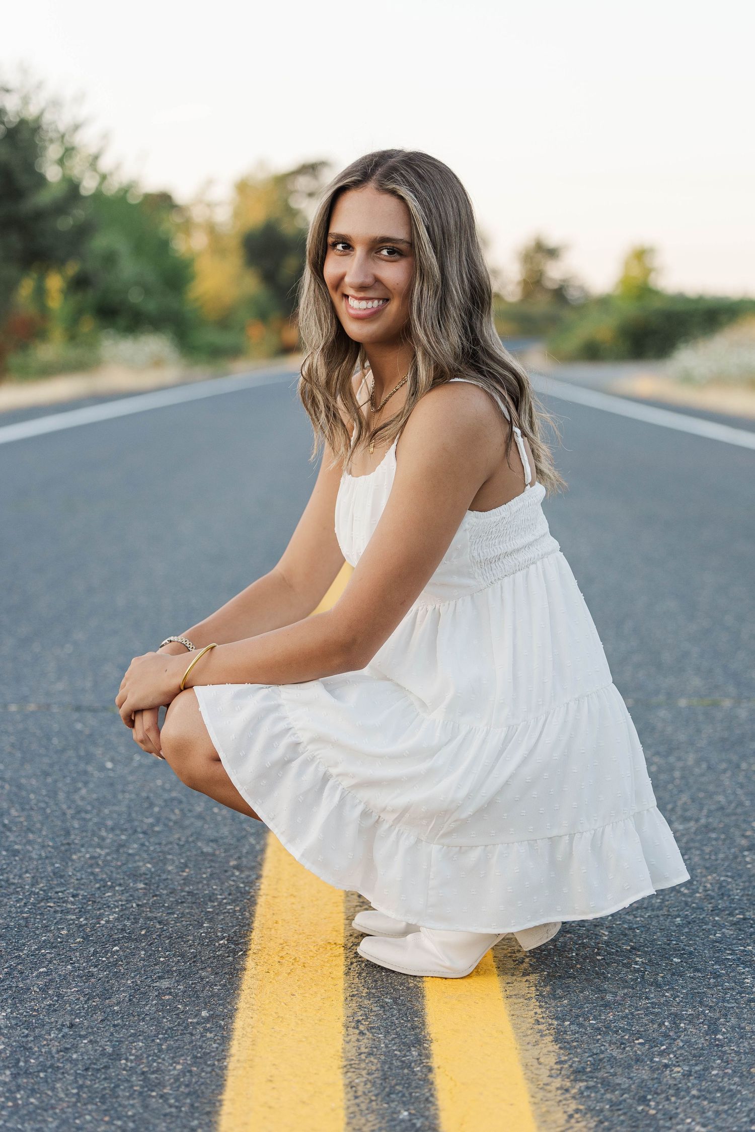 Smiling senior girl in white dress crouches on yellow road centerline at sunset for portrait photo.