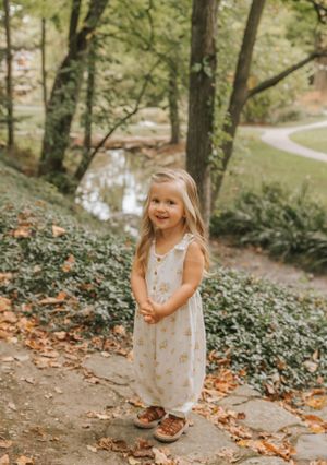 Small child in light dress explores wooded path with fallen autumn leaves.