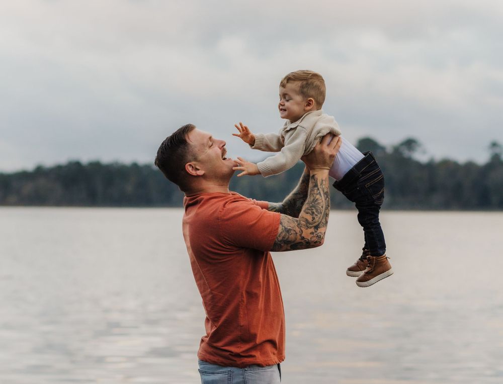 A playful moment between a parent and child lifting them up by a lake on an overcast day.