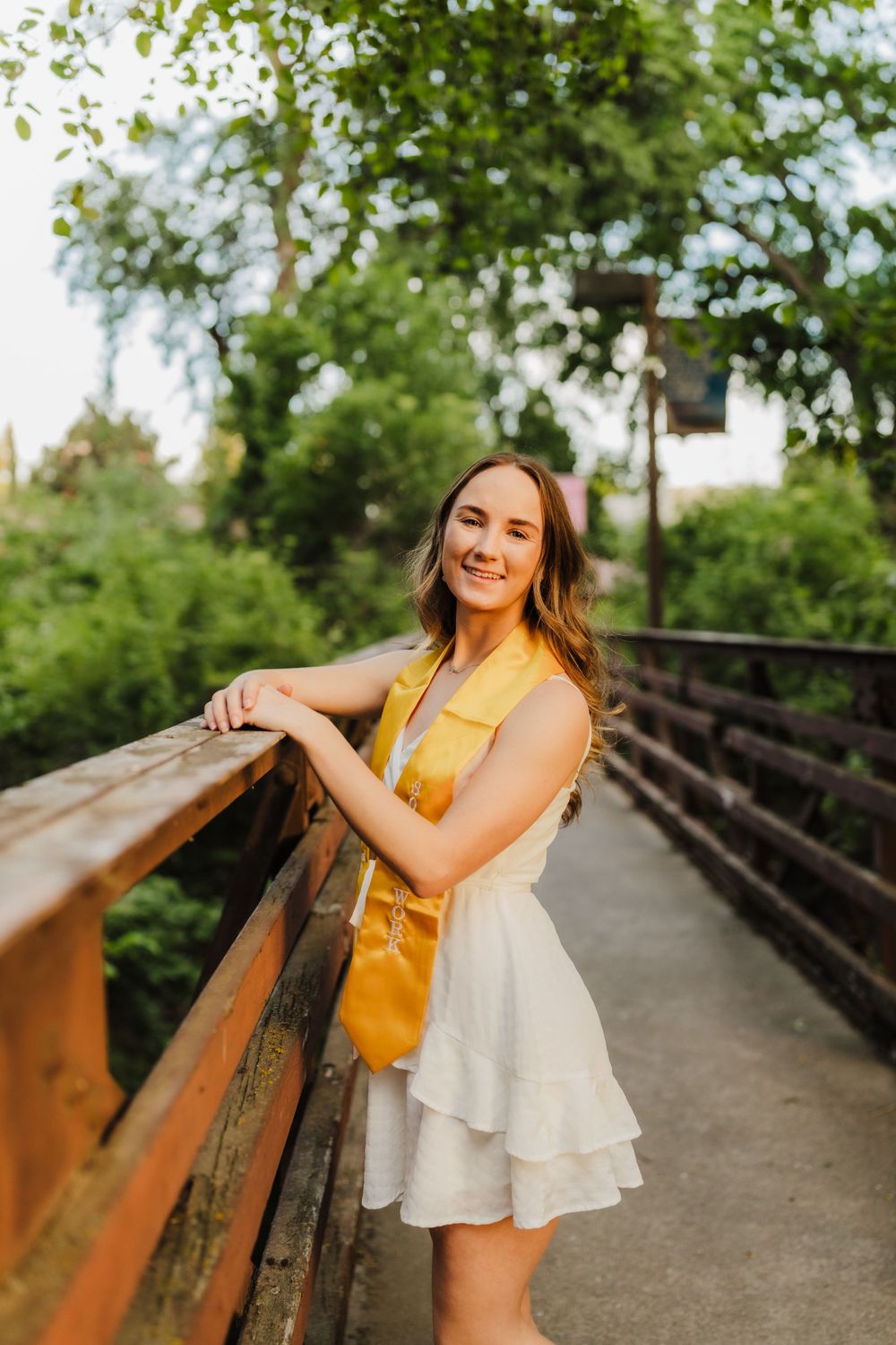 A figure in a yellow top and white layered dress poses on a rustic wooden bridge surrounded by greenery.