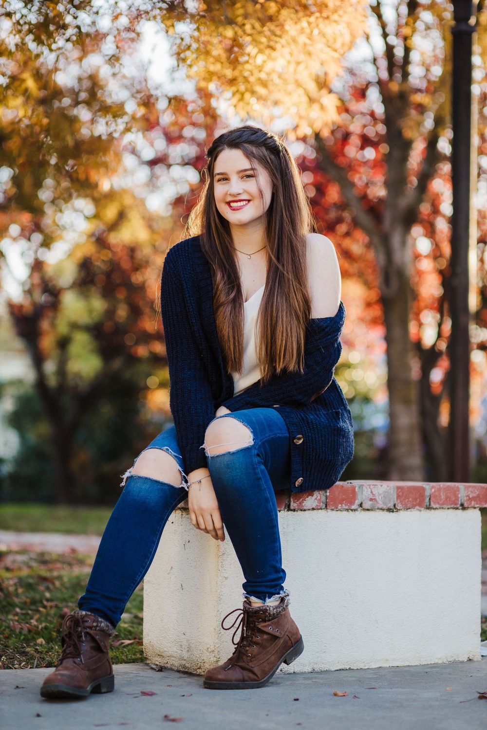 Woman in navy sweater and ripped jeans sitting on white wall during autumn with colorful fall foliage in background.