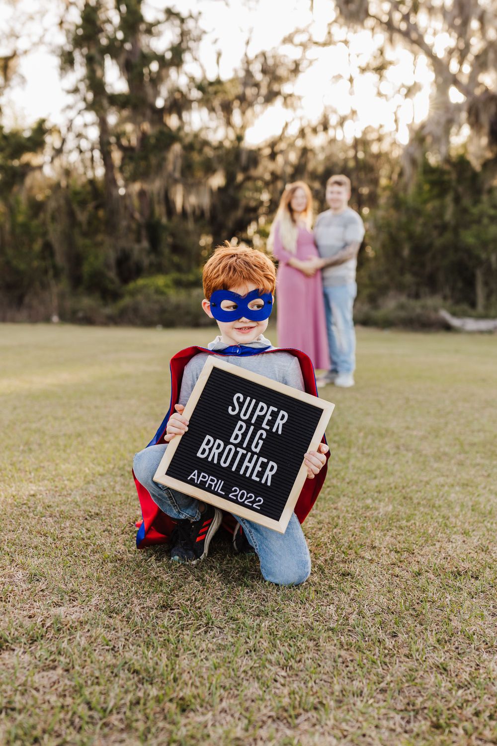 Child in superhero costume holding 'Super Big Brother' sign with family in background.