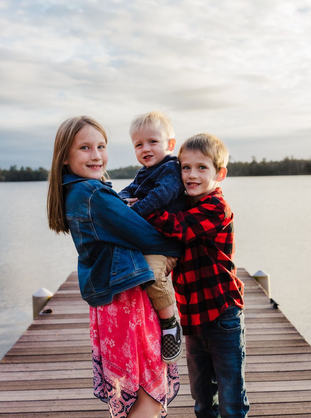 Three children pose together on a wooden dock overlooking a serene lake.
