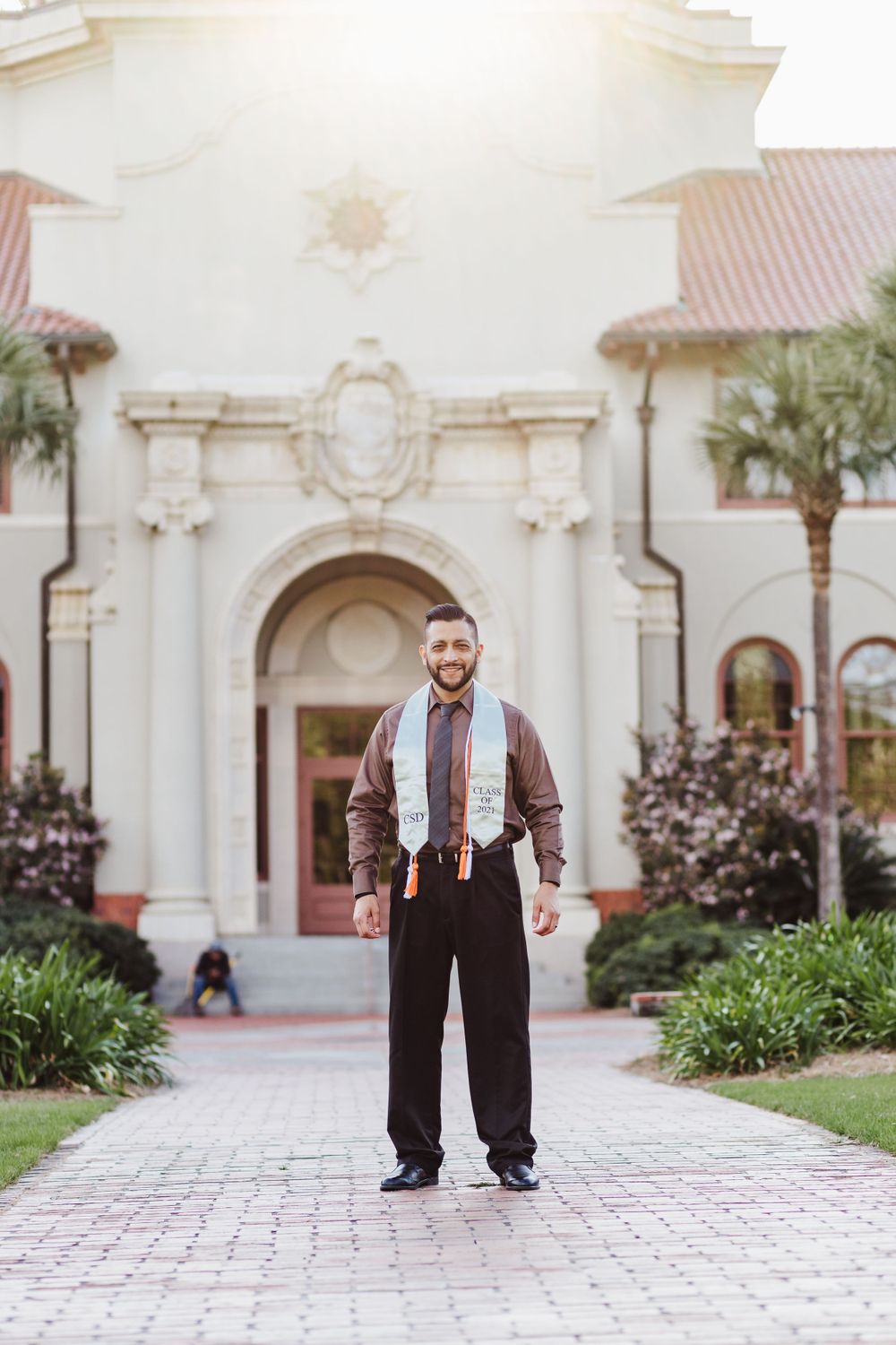 A person in vintage-style clothing with suspenders stands on a paved walkway in front of a Spanish colonial building.