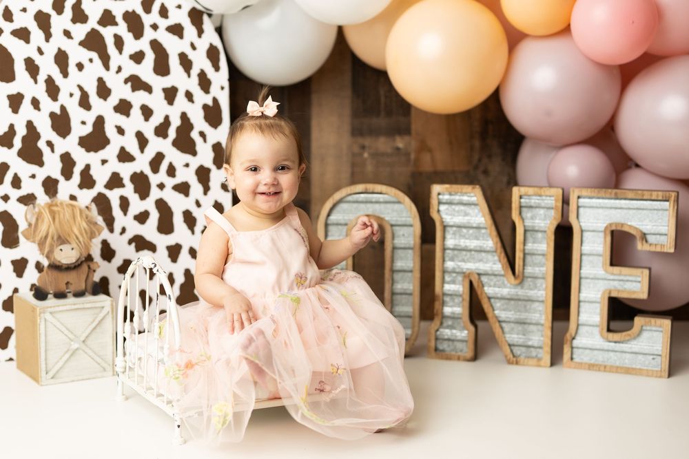 Baby girl in pink dress sits by ONE marquee letters with balloons and cow print backdrop for first birthday.