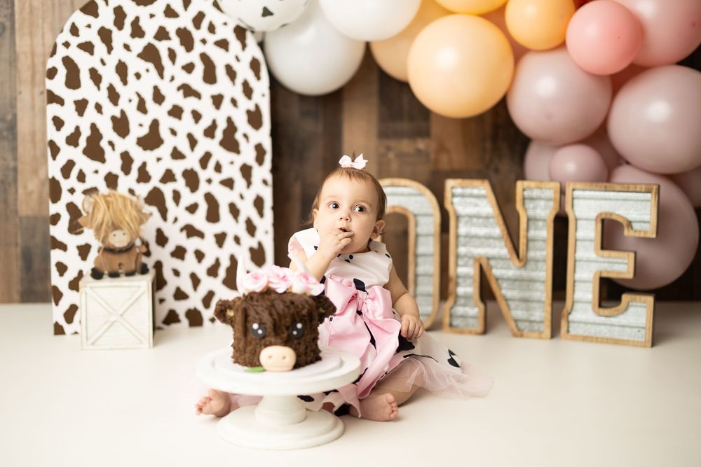 Baby girl celebrates first birthday smash cake with cow print backdrop and ONE marquee letters and balloon arch.