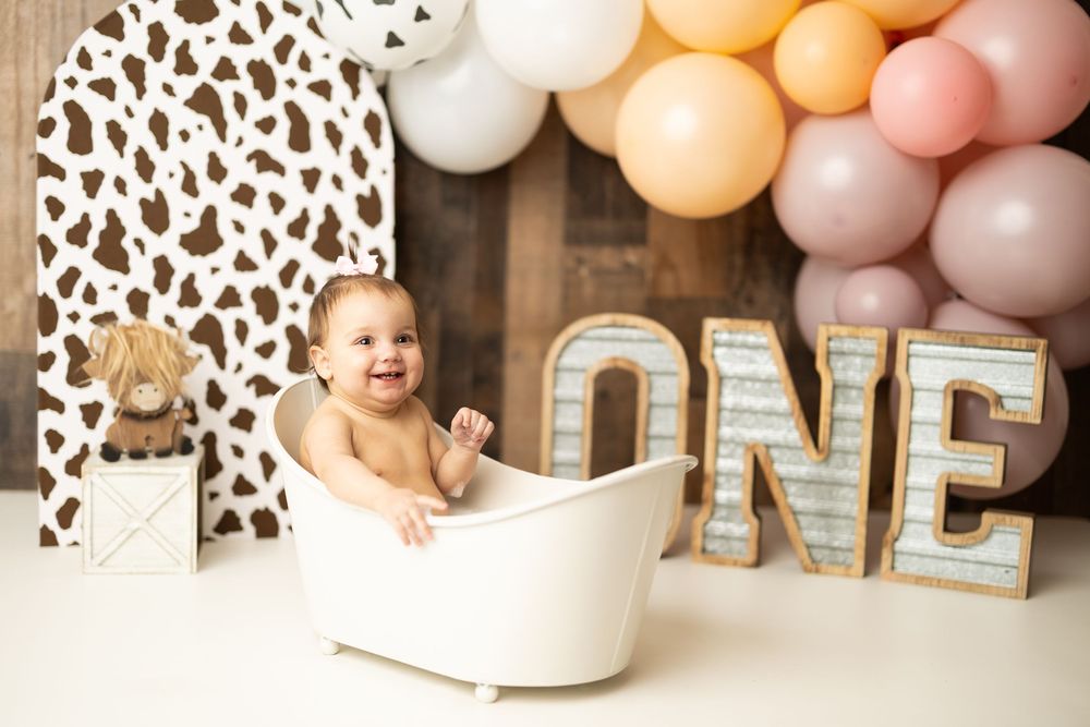 Smiling baby in white tub at first birthday cake smash with ONE sign, cow print backdrop, and balloon garland.