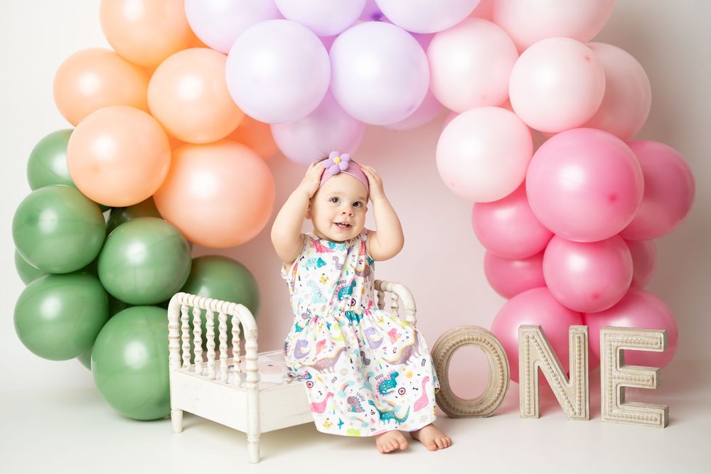 Baby girl celebrates first birthday with colorful balloon arch and ONE letters in pastel pink, green, and peach.