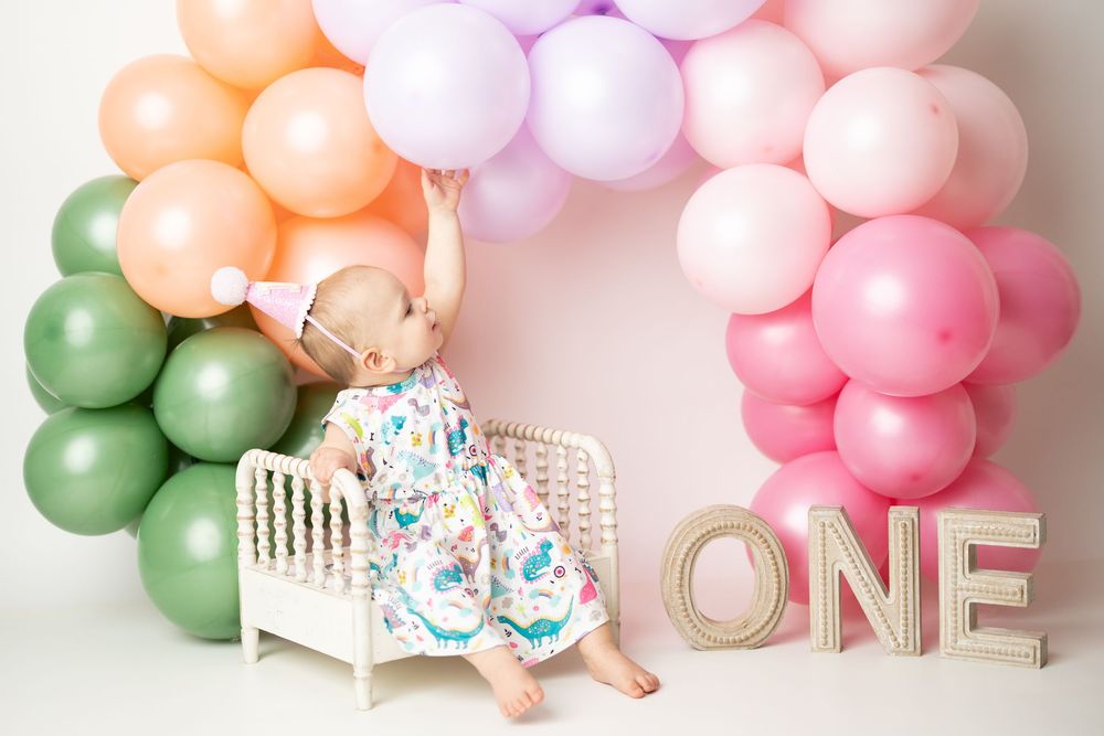 Baby in floral dress sits in mini crib under colorful balloon arch with ONE sign at first birthday party.
