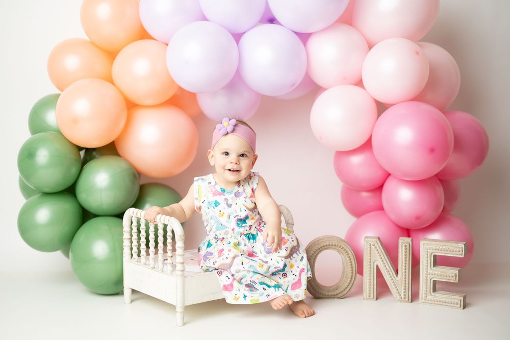Baby girl in floral dress sits by mini bed with colorful balloon arch and ONE sign at first birthday party.