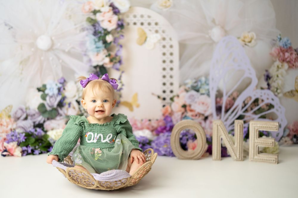 Baby girl in green outfit sits in basket celebrating first birthday with floral ONE backdrop decorations.