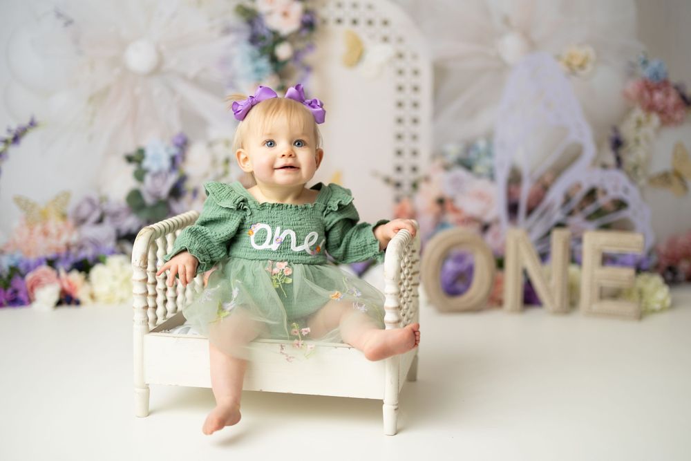 Baby girl in green \"one\" dress with purple bow sits on white bench for first birthday photo session.