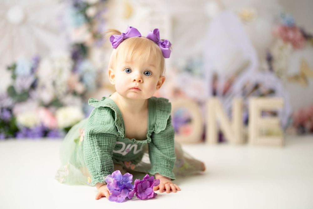 Baby girl in green dress with purple bow crawls near floral decorations and ONE sign for first birthday photo.