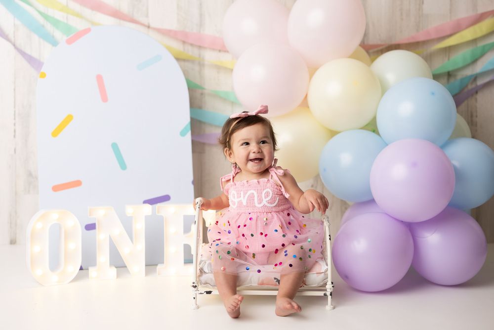 Baby girl in pink tutu sits smiling beside ONE sign surrounded by pastel balloons at first birthday cake smash photo shoot.